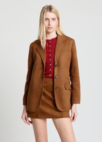 Blazer en daim marron avec une coupe ajustée, doté de deux poches avant et de boutons marron. Porté sur un haut boutonné bordeaux et une jupe.