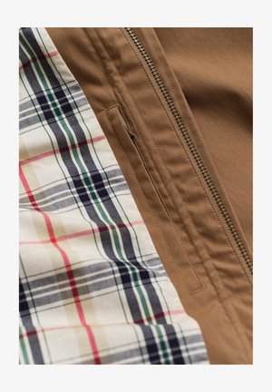 Brauner Jackenstoff mit einem Metallreißverschluss und ein kariertes Hemd in Beige, Marine, Grün und Rot unter der Jacke.