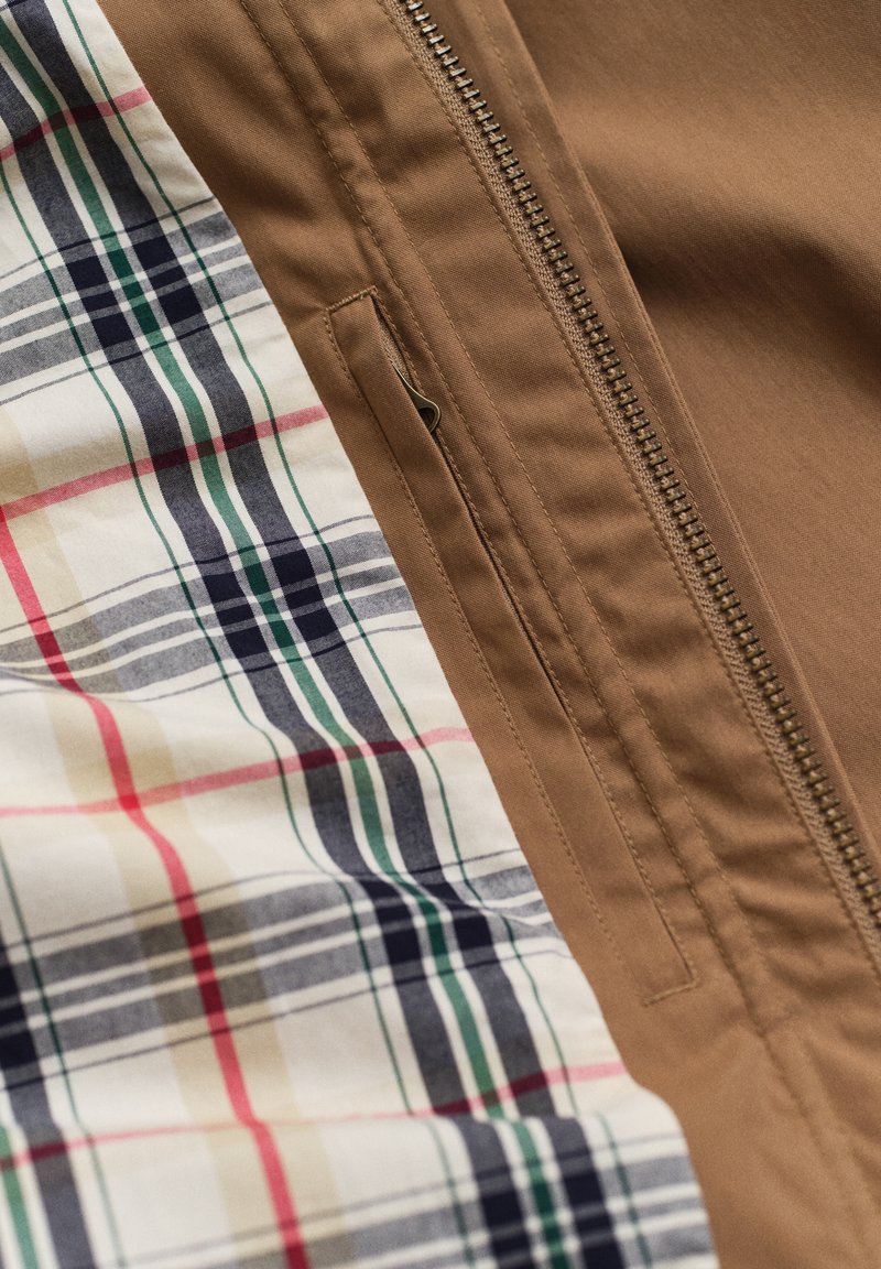 Tissu de veste marron avec une fermeture éclair en métal et une chemise à carreaux beige, marine, verte et rouge sous la veste.