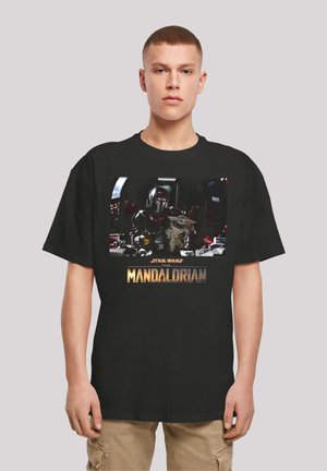Schwarzes T-Shirt mit einem bunten Grafikdesign eines Charakters in Rüstung, der ein kleines Wesen hält, mit dem Text "Star Wars The Mandalorian" darunter.