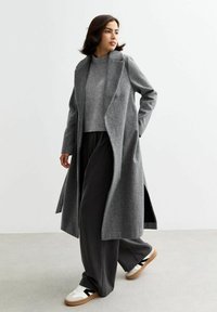 Manteau en chevrons gris au design ajusté, porté sur un pull gris et un pantalon noir ample, associé à des baskets blanches avec des accents noirs.