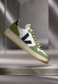Grønn og hvit sneaker med en teksturert semsket lær overdel, svarte detaljer, lysebeige såle og kremfargede snørebånd; minimalistisk design med logodetaljer.