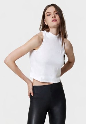 Donna che indossa un top corto bianco senza maniche e pantaloni in pelle neri a vita alta, in piedi con le mani sui fianchi contro uno sfondo semplice.