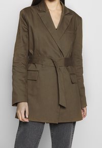 Femme portant un blazer ceinturé vert olive avec poches plaquées sur un jean gris foncé, se tenant contre un fond clair.