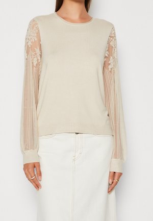 Beige villapaita, jossa on yksivärinen vartalo ja läpinäkyvät pitsihihat, joissa on kukka-embroidery ja pystyraitoja. Viimeistelty ribbireunuksilla.