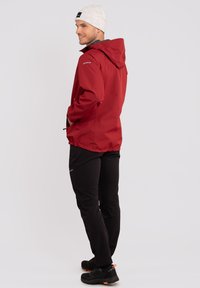 Rode waterdichte jas met een capuchon, elastische zoom en voorkruiszakken, gecombineerd met zwarte broek en zwarte wandelschoenen. Lichtgrijze beanie op het hoofd.