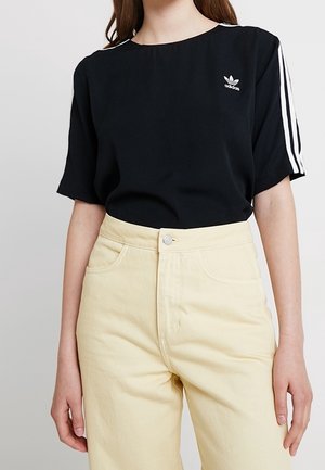 Frau trägt ein schwarzes Adidas-T-Shirt mit weißen Streifen an den Ärmeln und cremefarbene, hoch taillierte Hosen vor einem schlichten Hintergrund.