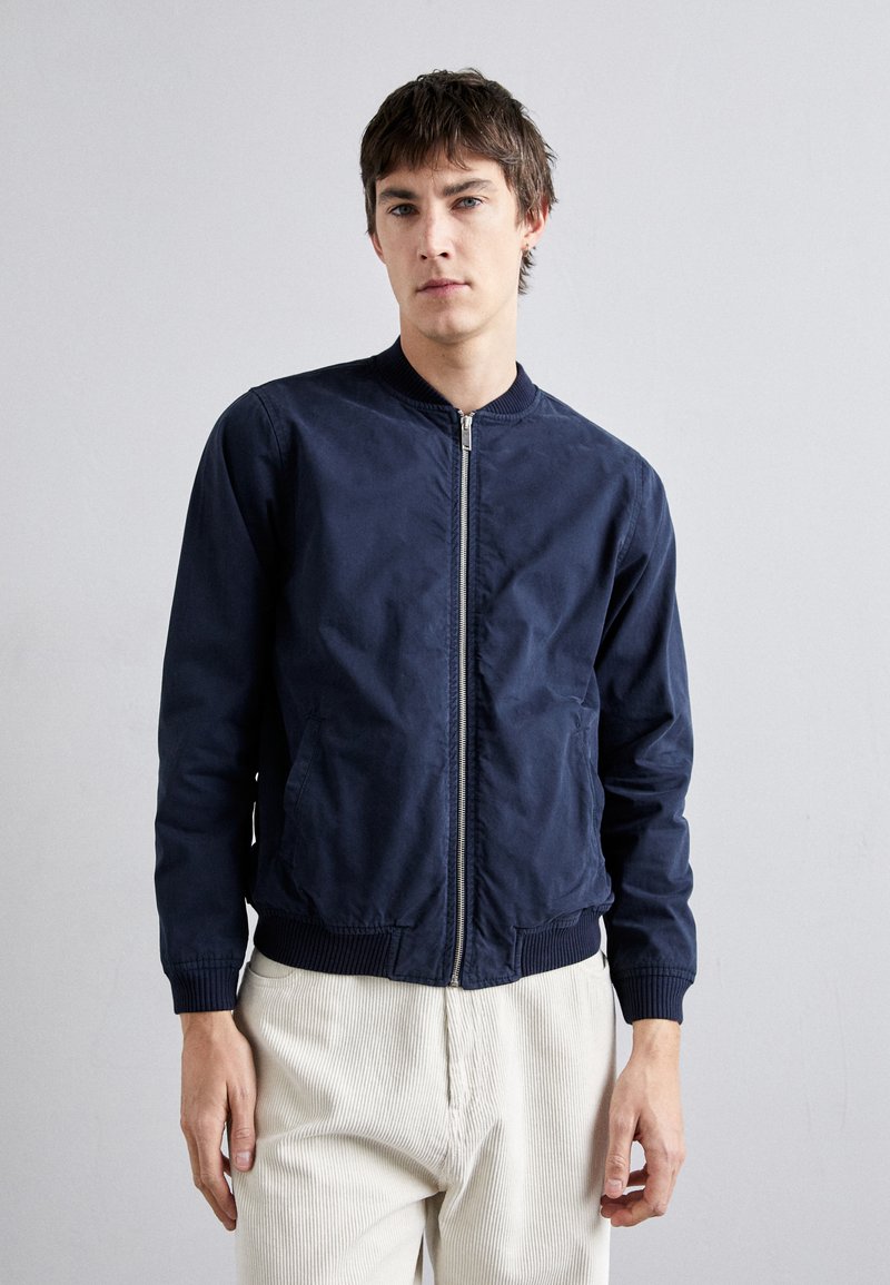 Chaqueta bomber navy en tejido ligero con puños y cuello acanalados, que cuenta con un cierre frontal y bolsillos laterales. Acabado texturizado en general.
