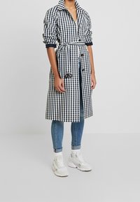 Rutig lång kappa i marinblått och vitt, med bälte, knäppning framtill och överdimensionerad krage. Bärs över jeans och sneakers.