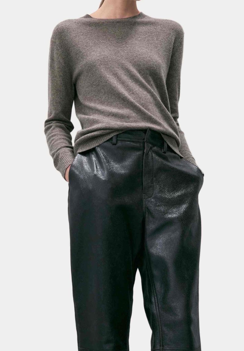 Femme portant un pull gris à manches longues rentré dans un pantalon en cuir noir brillant taille haute, les mains dans les poches.