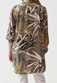 Persona che indossa una camicia a maniche lunghe, oversize, con le maniche arrotolate e un grande motivo floreale astratto in toni terrosi smorzati.