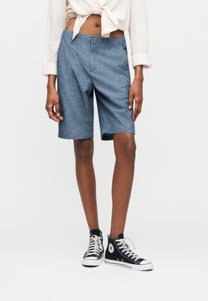 Osoba oblečená v svetlomodrých krátkych nohaviciach po kolená, bielej previazané košeli a čiernych vysokých teniskách na bielom pozadí.