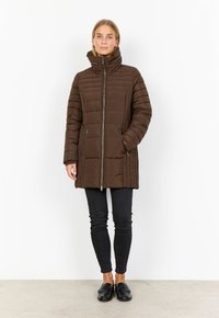 Brun pufferjacka med quiltad textur och hög krage, med en framzipper och två sidofickor, kombinerad med svarta smala jeans.