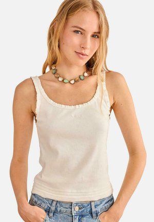 Mujer joven con top blanco sin mangas de canalé, collar multicolor de cuentas y vaqueros azules con las manos en los bolsillos.