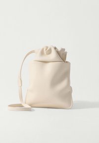 Lille beige læderpung med snørelukning og lang justerbar rem, stående oprejst på en hvid overflade.