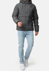 Man som bär en mörkgrå pufferjacka med huva, ljusblå jeans och vita sneakers, står och går framåt på en vit bakgrund.