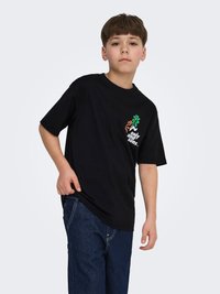 Pojke som bär en svart t-shirt med en färgglad grafik och text på bröstet, ihop med mörkblå jeans, stående mot en enfärgad bakgrund.