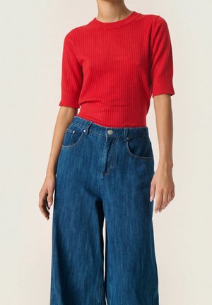 Personne portant un haut côtelé rouge vif à manches courtes, rentré dans un jean en denim bleu taille haute à jambes larges.