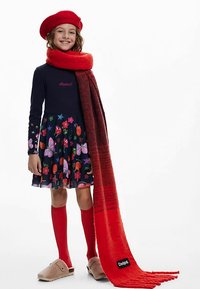 Czerwona beret i szalik, granatowa sukienka z kwiatowymi i motylkowymi wzorami, czerwone skarpety za kolano i beżowe chodaki. Miękkie tekstury i żywe kolory.