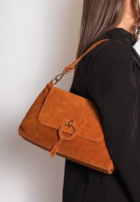 Bruine suède schoudertas met een afgeronde ringaccent, tassel-detail en een klep-sluiting. De schouderriem is verstelbaar.