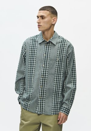 Homme aux cheveux courts portant une chemise verte rayée et à carreaux boutonnée et un pantalon beige, regardant vers sa gauche, debout devant un fond blanc.