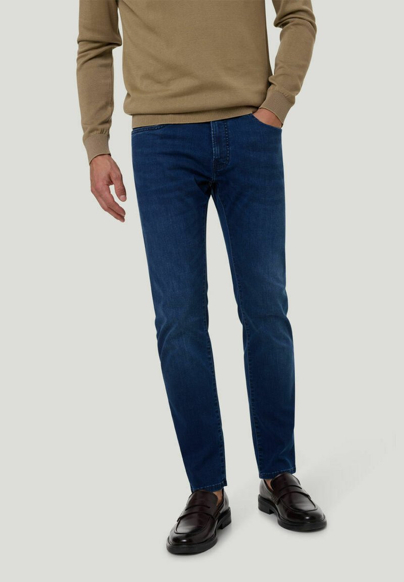 Jean en denim bleu foncé avec une coupe slim, associé à un pull en tricot beige et des mocassins marron foncé. Les jeans présentent une texture subtile et aucune usure visible.