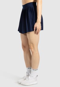 Marineblauwe geplooide sportrok met een brede tailleband, gemaakt van rekbaar materiaal. Gestyled met witte sportschoenen en sokken.