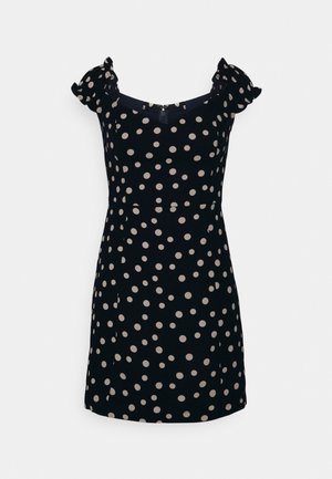 Musta pilkullinen mekko lyhyillä puffihiusilla, pyöreällä kaula-aukolla ja tiukalla siluetilla. Takana on vetoketjukiinnitys.