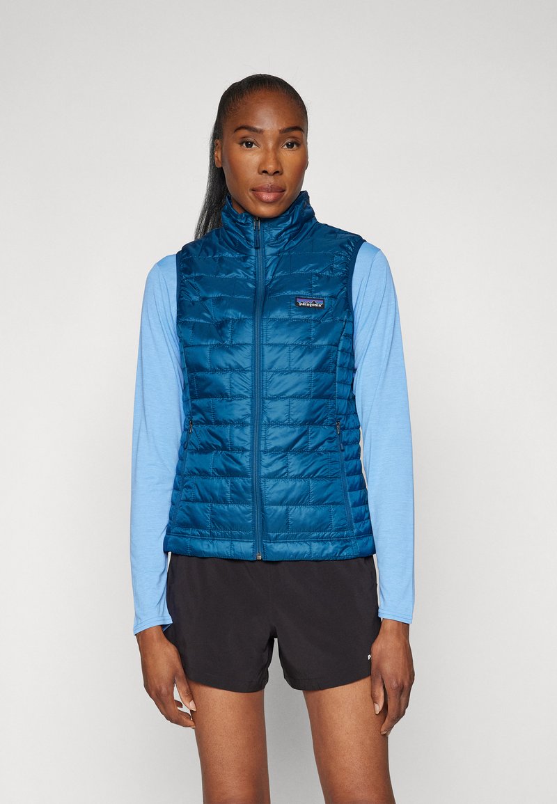 Gilet matelassé bleu avec un col haut, fermeture éclair sur le devant et poches latérales, porté sur une chemise à manches longues bleu clair, sur un fond neutre.