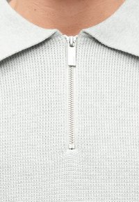 Sweater texturizado cinza-claro com colarinho e fecho frontal com zíper, apresentando uma aba metálica. Padrão de malha waffle por todo o lado.