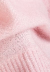 Ljust rosa stickad tyg med en mjuk, fluffig textur; har ribbad detalj längs kanten. Det övergripande intrycket är mysigt och lätt.