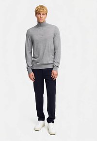 Hellgrauer Langarm-Pullover mit Reißverschluss-Kragen, kombiniert mit dunkelblauen Hosen und weißen Turnschuhen. Glatte Textur, tailliertes Design.