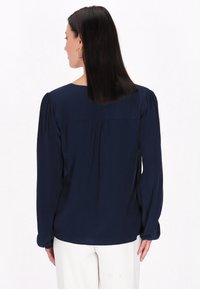 Marineblå bluse med lange pufærmer, rygplissering og rund halsudskæring. Fremstillet af et glat, let materiale.