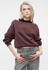 Jauna moteris su šviesiais plaukais, vilkinti laisvą bordo spalvos trumpą džemperį ant juodos palaidinės ir pilkų aukšto liemens džinsų, rankos poilsiauja ant klubų.