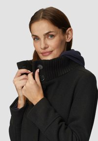 Zwarte jas met een geribbelde kraag, voorzien van grote zwarte knopen. De stof heeft een gestructureerde uitstraling. Gelaagd met een marineblauw hoodie.