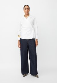 Blusa blanca con mangas largas y detalles dorados en los puños, combinada con pantalones vaqueros de pierna ancha en azul oscuro y zapatos de tacón de diseño.