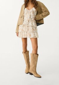 Bottes de cowboy en daim beige avec un bout pointu et des coutures décoratives, portées avec une robe florale à volants et une veste marron clair.