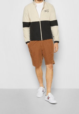 Veste en polaire beige et noire avec une fermeture éclair à l'avant ; portée avec un short en velours côtelé marron et des baskets blanches avec des accents rouges et bleus.