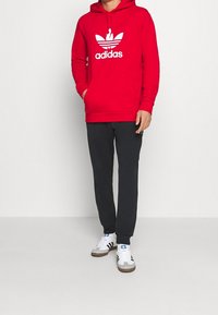Felpa rossa con logo Adidas bianco, tasca a canguro, abbinata a pantaloni della tuta neri e sneakers grigie con strisce nere e accenti blu.