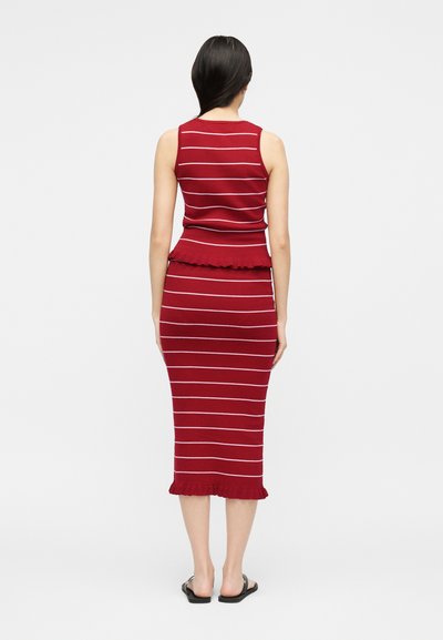 Γυναίκα που φοράει κόκκινο αμάνικο φόρεμα με λεπτές λευκές οριζόντιες ρίγες και βολάν στο τελείωμα, στεκόμενη πλάτη σε μονόχρωμο φόντο.
