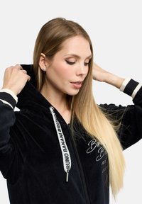 Svart velour zip-up hoodie med vita dragsnören, broderad text och ribbad mudd. Modellen har långt hår och justerar huvan.