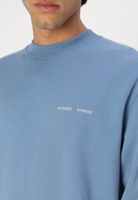 Blue sweatshirt made of soft fabric, featuring a crew neckline and two embroidered labels reading "SAMSØE SAMSØE" on the chest.