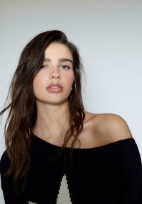 Schwarzer Strickpullover mit schulterfreiem Schnitt und großem weißen Sternenmuster. Das Modell hat lange, dunkle Haare und einen natürlichen Make-up-Look vor einem neutralen Hintergrund.