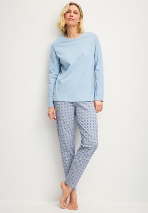 Vrouw draagt lichtblauw shirt met lange mouwen en een pyjamabroek met patroon, staat blootsvoets tegen een effen lichte muur.