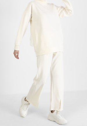 Person iført løstsiddende cremefarvede bukser med sidelommer, matchende oversize sweatshirt og hvide chunky sneakers, poserende mod hvid baggrund.