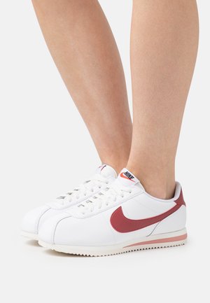 Nohy obuté v bílých teniskách Nike Cortez s červenými logy a patami, stojící na bílém povrchu.