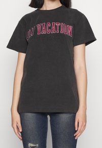 Osoba nosząca ciemnoszary t-shirt z krótkim rękawem z czerwonym i białym napisem "NA WAKACJACH" oraz wyblakłe niebieskie dżinsy z przetarciem na lewym kolanie.