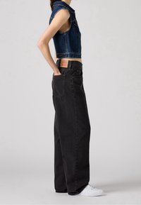 Personne portant un gilet en denim bleu sans manches et un jean noir ample avec des baskets blanches, debout de profil avec la main dans la poche arrière.