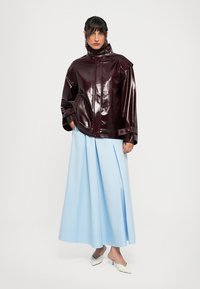 Femeie îmbrăcată într-o jachetă oversized strălucitoare de culoare burgundă închisă, o fustă plisată albă-bleu și pantofi cu vârf ascuțit albi, stând în fața unui fundal alb.