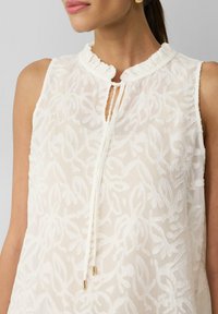 Witte mouwloze blouse met een textuur van bloemmotieven, een volantskleur, en een keyhole sluiting met strikdetails en gouden accenten.